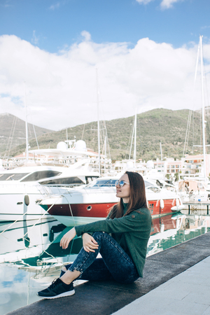 A beautiful young girl in sunglasses is sitting next to the yachts in Porto Montenegro in Tivat in Montenegro resting and looking at the yachts.の写真素材