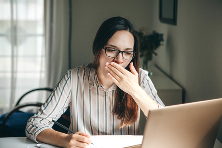 A girl works at a computer in a home office or a student at home teaches lessons. She is tired, yawns and wants to sleep or is bored.の写真素材