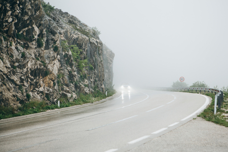 View of the mountain road on which the car is driving in foggy and rainy weather. The car is in motion and it has its headlights turned on.の写真素材