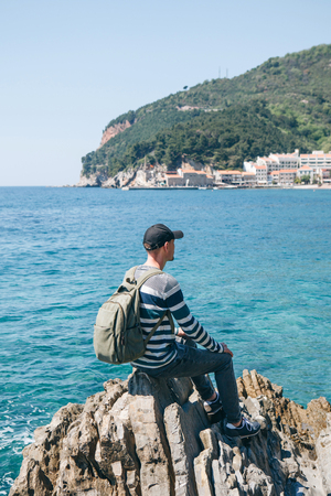 A tourist with a backpack alone sits on a cliff by the sea and looks into the distance at the view of Petrovac in Montenegro.の写真素材