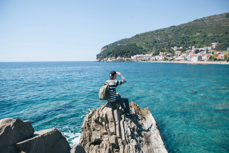 A tourist with a backpack alone sits on a cliff by the sea and looks into the distance at the view of Petrovac in Montenegro.の写真素材