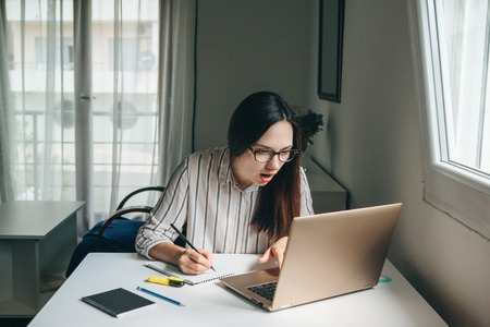 A young beautiful student girl does homework or works in a home office. She is surprised by the news or she has found an unexpected solution.の写真素材