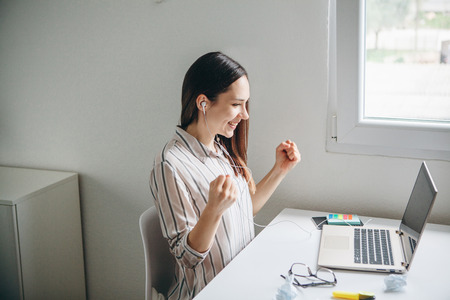 Girl in headphones working at a laptop in the home office. She found a solution or saw good news or an idea and rejoices.の写真素材