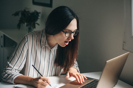 A young beautiful student girl does homework or works in a home office. She is surprised by the news or she has found an unexpected solution.の写真素材
