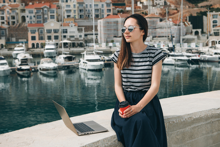 Portrait of a positive young woman working on a laptop and drinking coffee outdoors. Freelancer or blogger or other.の写真素材