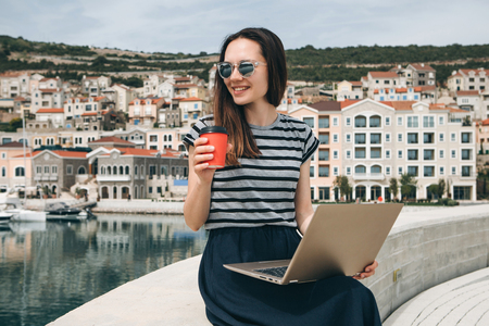 Portrait of a positive smiling young woman working on a laptop and drinking coffee outdoors. Freelancer or blogger or other.の写真素材