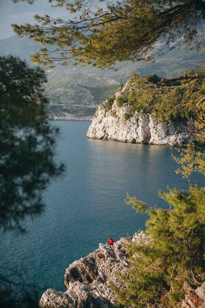 Beautiful view through tree branches to the sea and natural landscape. Among the rocky shores of an unrecognizable man fishing.の写真素材