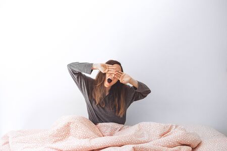 The girl is in bed and yawns. She covers her mouth with her hand. Early morning and she is sleepy.の写真素材