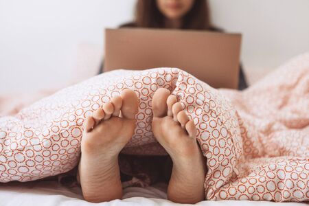 Selective focus on the legs. Young adult beautiful girl enjoys a laptop in bed. She works either on the Internet or watching a movie.の写真素材