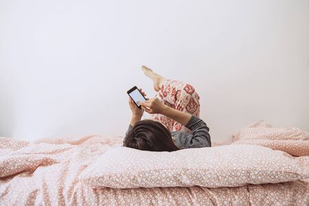 The girl lies in bed and listens to music or a podcast. The girl uses her cell phone.の写真素材