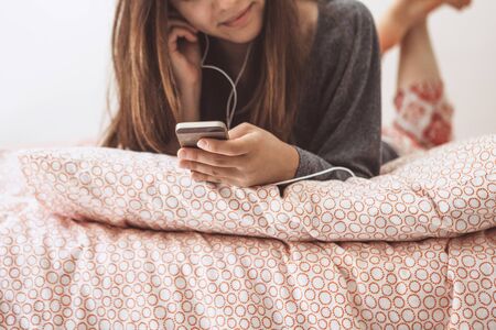 The girl lies in bed and listens to music or a podcast. The girl uses her cell phone.の写真素材
