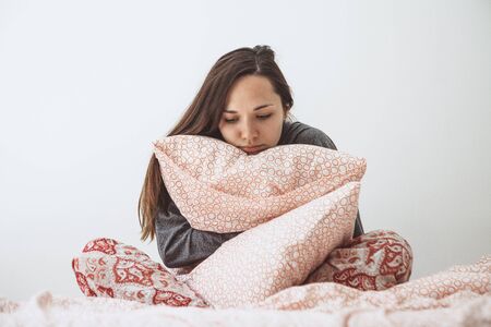 Sad or upset girl sitting in bed with a pillow. She is ill, lonely or in a bad mood or other problems.の写真素材
