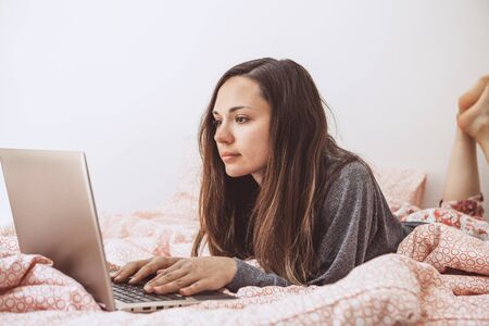 Young adult beautiful girl enjoys a laptop in bed. She works either on the Internet or watching a movie.の写真素材