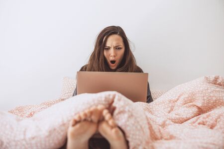 The girl uses the computer in bed. She saw something and was surprised or shocked.の写真素材