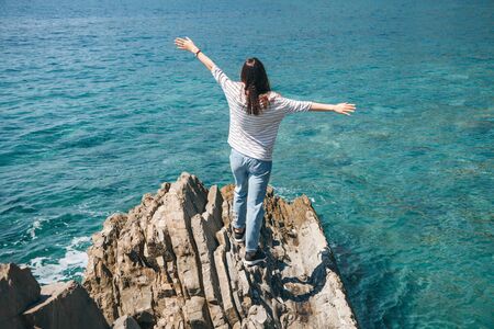 A girl stands on a rocky shore by the sea and raises her hands up showing how happy and free she is. Digital detox.の写真素材
