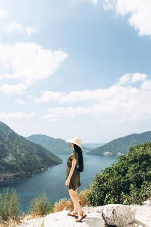 A girl in a hat on the background of the sea and mountains admires a beautiful view of the natural landscape in Montenegro.の写真素材