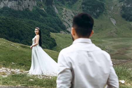 The groom looks or admires his beautiful bride in a white dress, which stands against the backdrop of a mountain landscape. Outdoor weddingの写真素材