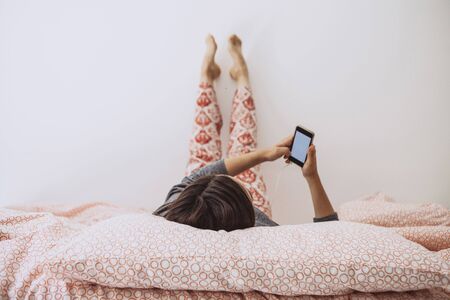 The girl lies in bed and listens to music or a podcast. The girl uses her cell phone.の写真素材