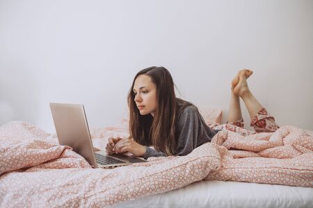 Young adult beautiful girl enjoys a laptop in bed. She works either on the Internet or watching a movie.の写真素材