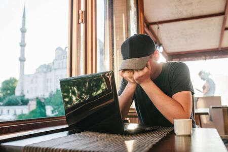Man in cafe sits at table with computer near window and hidden face by handsの写真素材