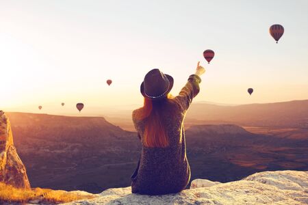 A woman in solitude sits on top of a mountain and admires the flight of hot air balloons in Cappadocia in Turkey. Digital detox and soul searchの写真素材