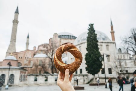 A person holds a traditional Turkish pretzel Simit against the background of the landmarks of Hagia...