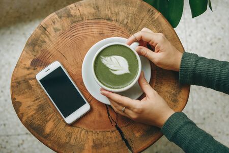 View from above. Girl holds in her hand a cup with fragrant fresh and healthy green matcha latte tea on a wooden table. Nearby lies a cell phone.の写真素材