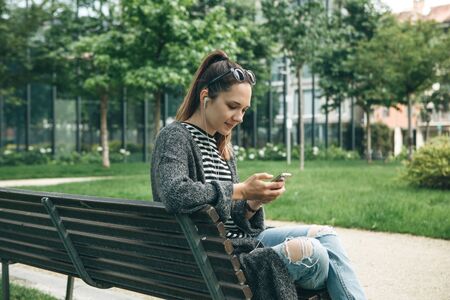 A young beautiful girl sits on a bench in a city park and plays music or a podcast or learns from audio lessons.の写真素材