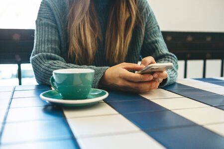 A girl drinks hot tea or coffee in a cafe and uses a cell phone or mobile phone.