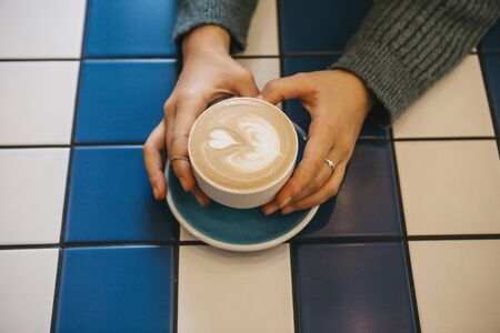 View from above. Girl holds in her hand a cup with cappuccino coffee on a table of blue and white ceramic tiles.の写真素材