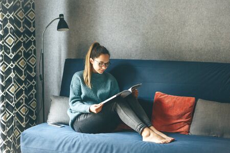 A girl reads an entertainment or educational magazine while sitting on a sofa in a cozy home environment.の写真素材