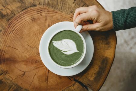 View from above. Girl holds in her hand a cup with fragrant fresh and healthy green matcha latte tea on a wooden table.の写真素材