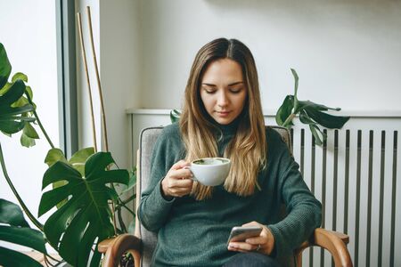 Girl drinks a hot morning drink and uses a cell phone.の写真素材