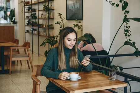 The girl uses a cell phone and drinks coffee. She has a meeting or she drinks a morning cappuccino or flat white.の写真素材