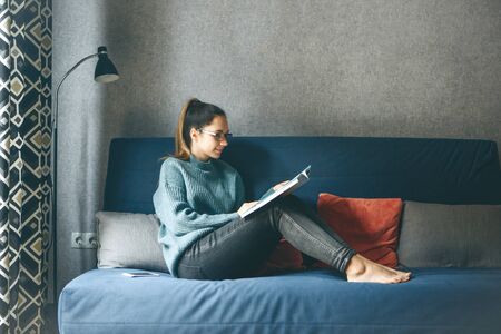 A girl reads an entertainment or educational magazine while sitting on a sofa in a cozy home environment.の写真素材