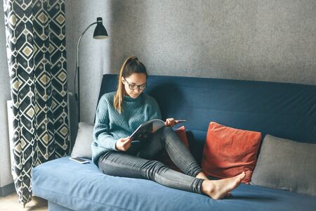 A girl reads an entertainment or educational magazine while sitting on a sofa in a cozy home environment.の写真素材