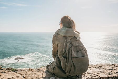Girl tourist with a backpack in solitude sits and looks at the seaの写真素材