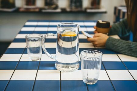 A decanter and a glass of water with lemon in the foreground. In the background, the girl uses a mobile phone. She has a meeting or she is resting and drinking coffee.の写真素材