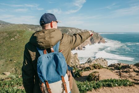 A tourist with a backpack shows his hand into the distance towards the sea or ocean.の写真素材