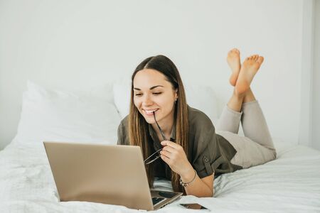 Positive girl student lies in bed and uses a laptop. She is reading from the screen, or she is watching a video or training video course or chatting online.の写真素材
