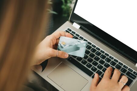 A girl holds a credit card or bank card in her hand next to a laptop. Laptop with white blank screen...