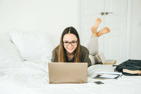 Positive girl student lies in bed and uses a laptop. She is watching a video or she is using video chat or online surfing on the Internet.の写真素材