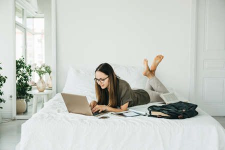 Positive girl student lies in bed and uses a laptop. She is texting or surfing online on the Internet.の写真素材