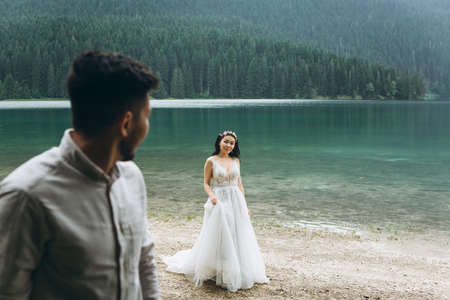 Beautiful young Kazakh bride in a white dress is walking on the background of the lake. The groom is looking at her.の写真素材