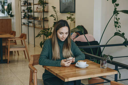 The girl uses a cell phone and drinks coffee. She has a meeting or she drinks a morning cappuccino or flat white.の写真素材