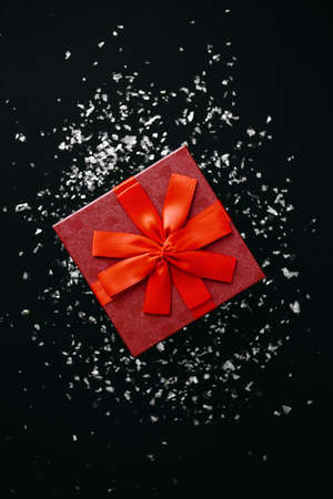 Red box with a gift on a dark background with many scattered sparkles or confetti nearby. Festive concept.の写真素材