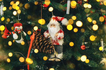 Close up of Santa Claus Christmas toy on the Christmas tree. Nearby is a snowman toy and a pine cone. There are lights around.の写真素材
