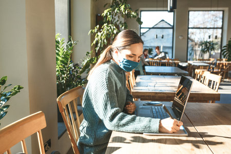 A girl in a protective mask uses a laptop and writes in a notebook.の写真素材