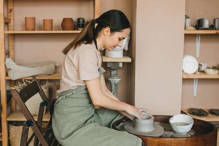 Young woman pottery maker works in clay studio on pottery wheel against shelves with vases and pots. Creative people concept.の写真素材