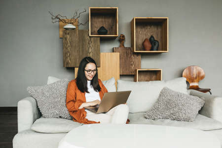 A beautiful European young woman using a laptop for remote work from home. A business woman in casual clothes conducts a video call, engages in e-mail, communicates online with colleagues, sends and responds to emails, develops software while sitting at home on the couch.の写真素材
